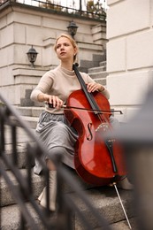 Photo of Beautiful young woman playing cello on stairs outdoors. Classic musical instrument