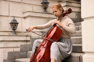 Photo of Beautiful young woman playing cello on stairs outdoors. Classic musical instrument