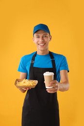 Fast-food worker holding paper container with fries and cup on orange background