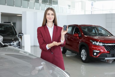 Photo of Happy saleswoman holding key near new silver car in salon