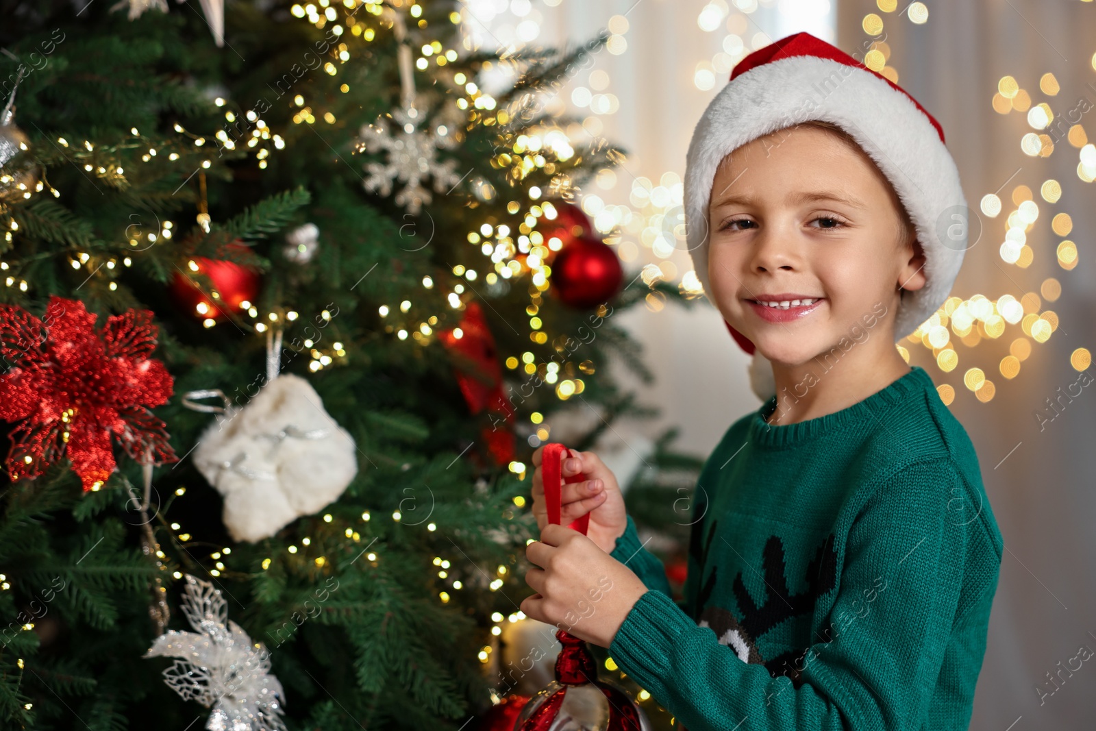 Photo of Cute little boy in Santa hat decorating Christmas tree at home