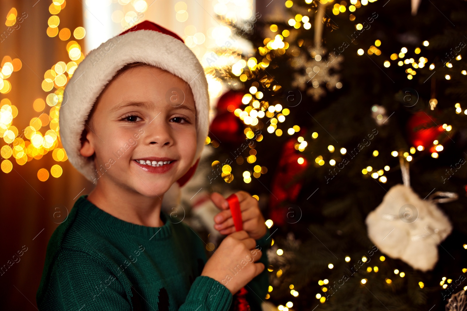 Photo of Cute little boy in Santa hat decorating Christmas tree at home