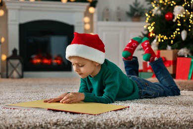 Cute little boy with Santa hat reading book on rug in room decorated for Christmas