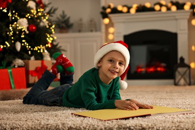 Photo of Cute little boy with Santa hat reading book on rug in room decorated for Christmas