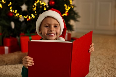 Photo of Cute little boy with Santa hat reading book in room decorated for Christmas