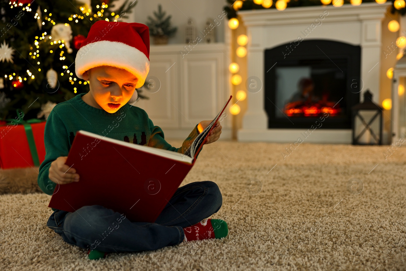 Photo of Cute little boy with Santa hat reading book on rug in room decorated for Christmas, space for text