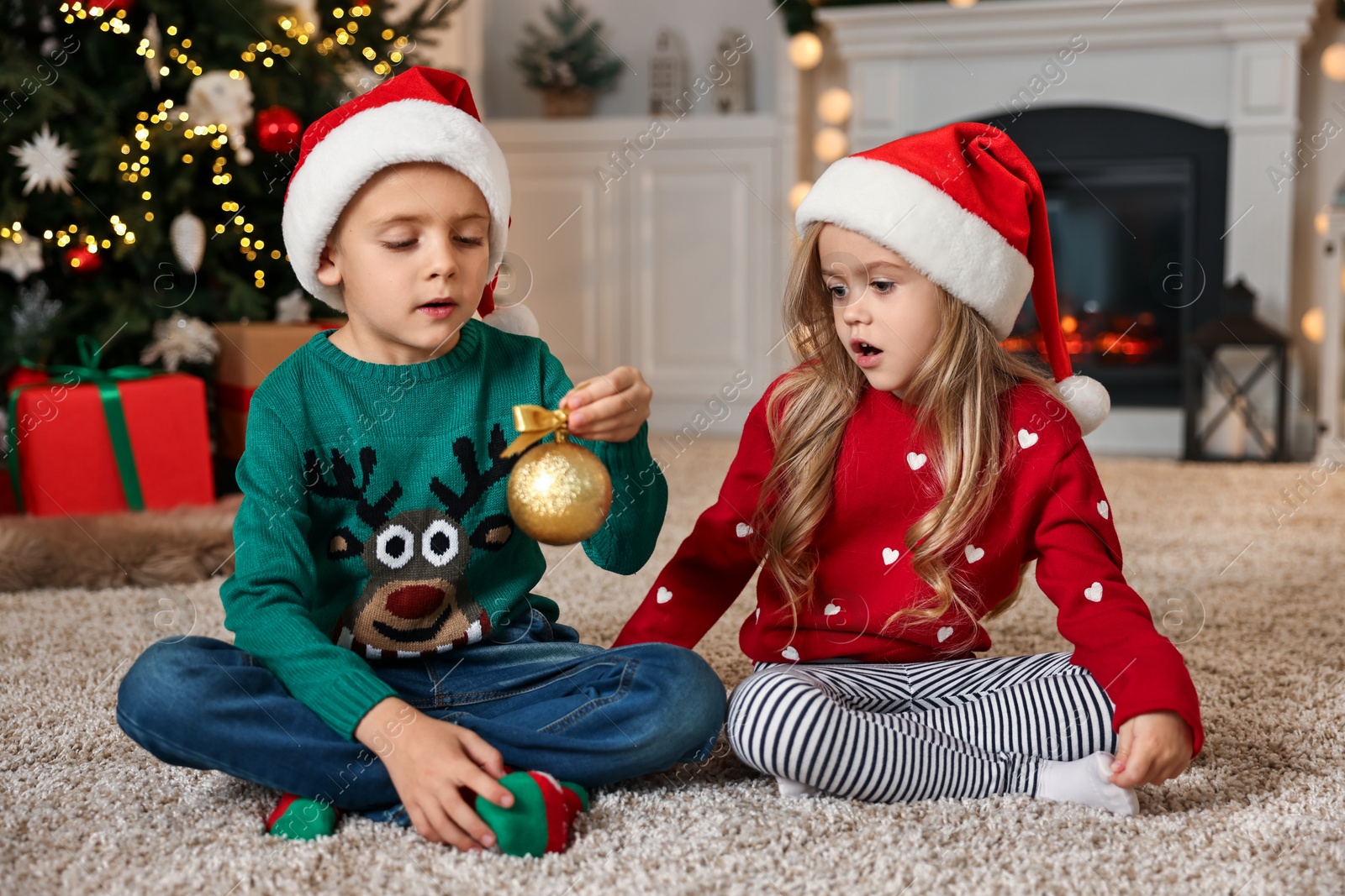 Photo of Little kids in Santa hats with Christmas ball on floor at home