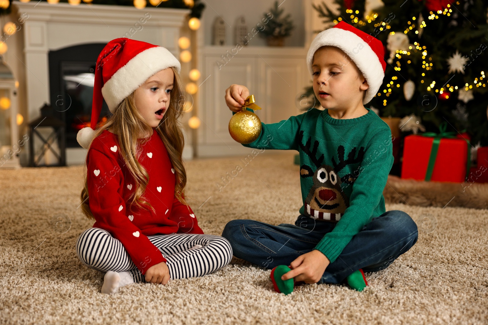 Photo of Little kids in Santa hats with Christmas ball on floor at home