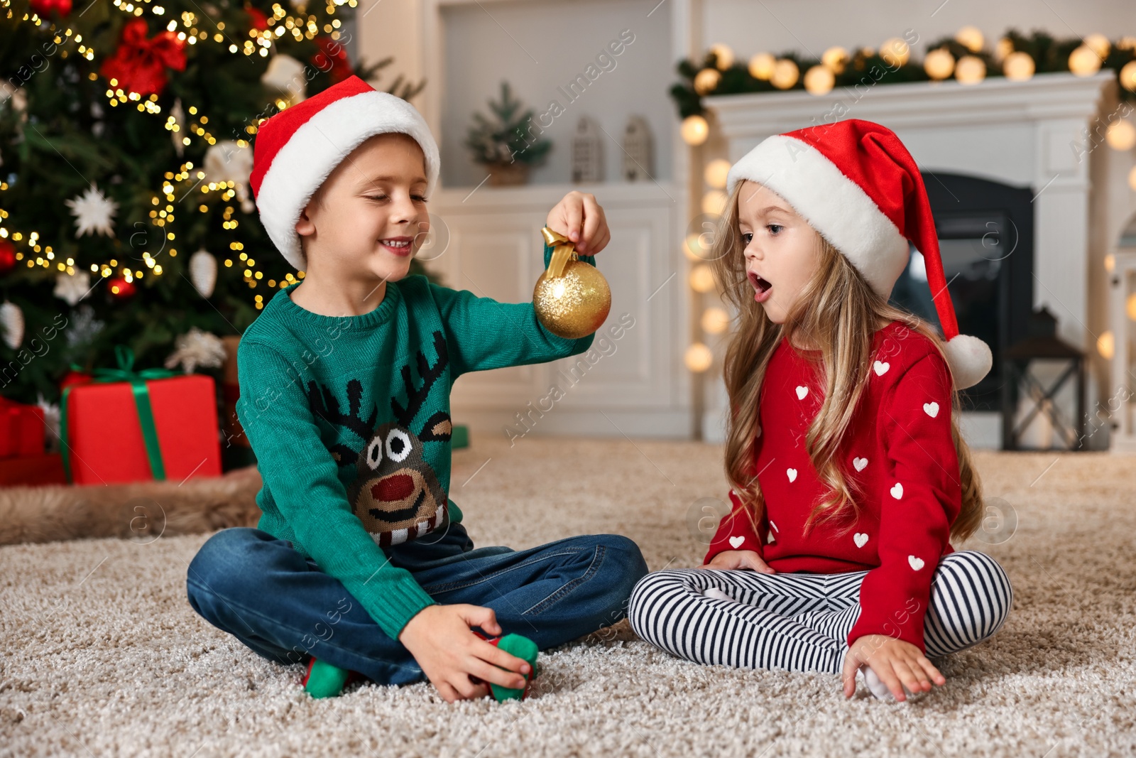 Photo of Little kids in Santa hats with Christmas ball on floor at home