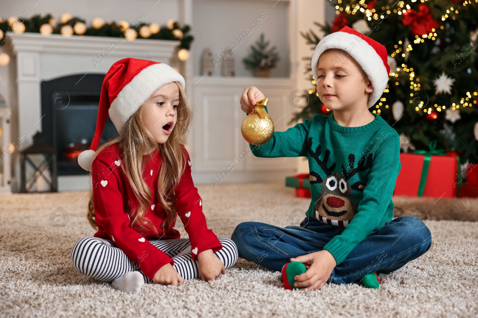 Photo of Little kids in Santa hats with Christmas ball on floor at home