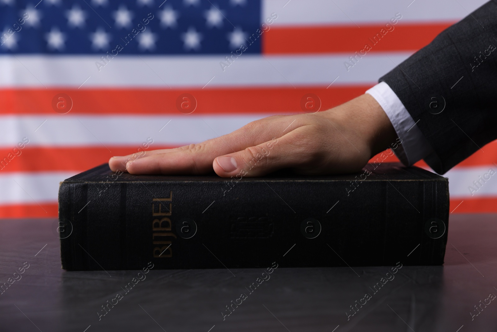 Photo of Man taking oath with his hand on Bible at black table against flag of USA, closeup