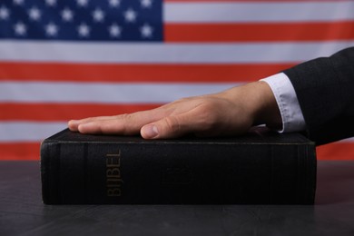 Photo of Man taking oath with his hand on Bible at black table against flag of USA, closeup