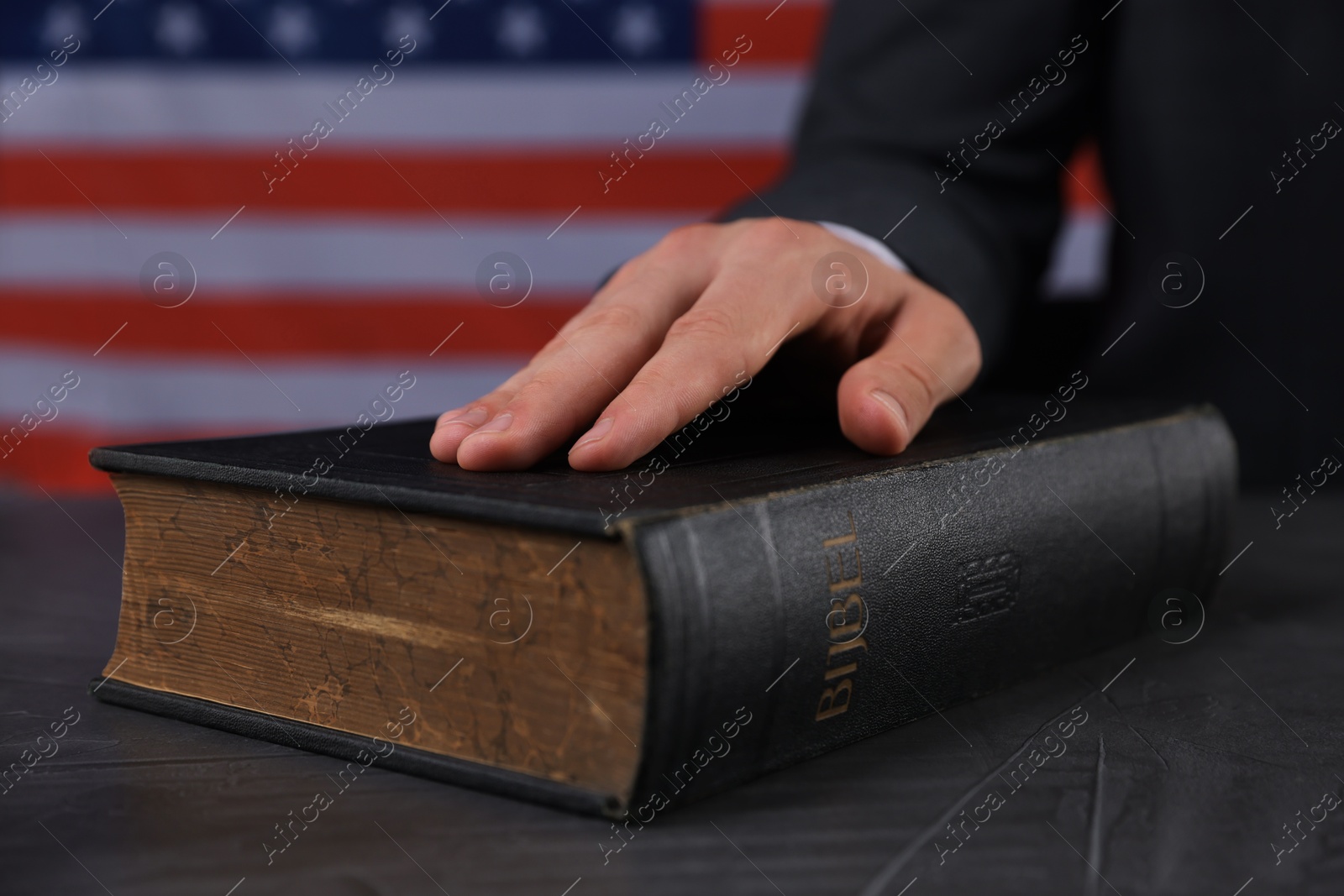 Photo of Man taking oath with his hand on Bible at black table against flag of USA, closeup