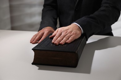 Photo of Man taking oath with his hand on Bible at white table, closeup