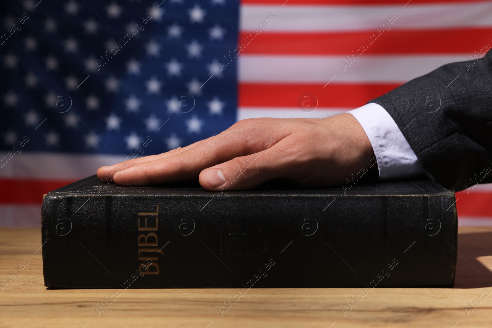 Photo of Man taking oath with his hand on Bible at wooden table against flag of USA, closeup