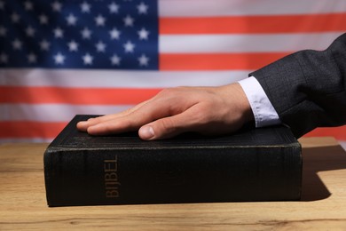 Photo of Man taking oath with his hand on Bible at wooden table against flag of USA, closeup