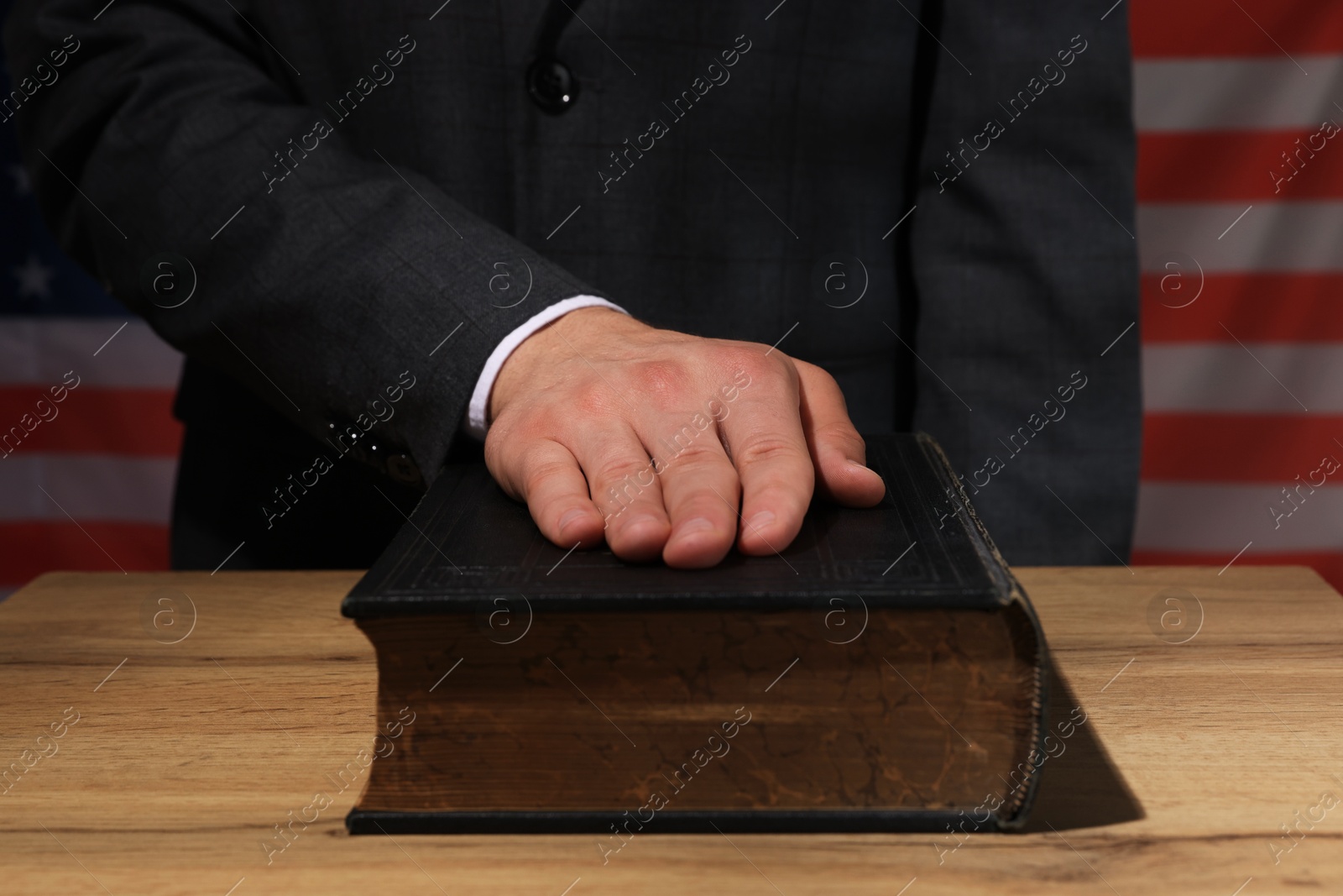 Photo of Man taking oath with his hand on Bible at wooden table against flag of USA, closeup