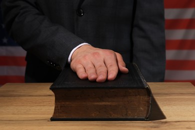 Photo of Man taking oath with his hand on Bible at wooden table against flag of USA, closeup