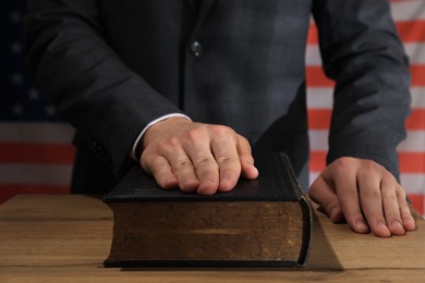 Photo of Man taking oath with his hand on Bible at wooden table against flag of USA, closeup