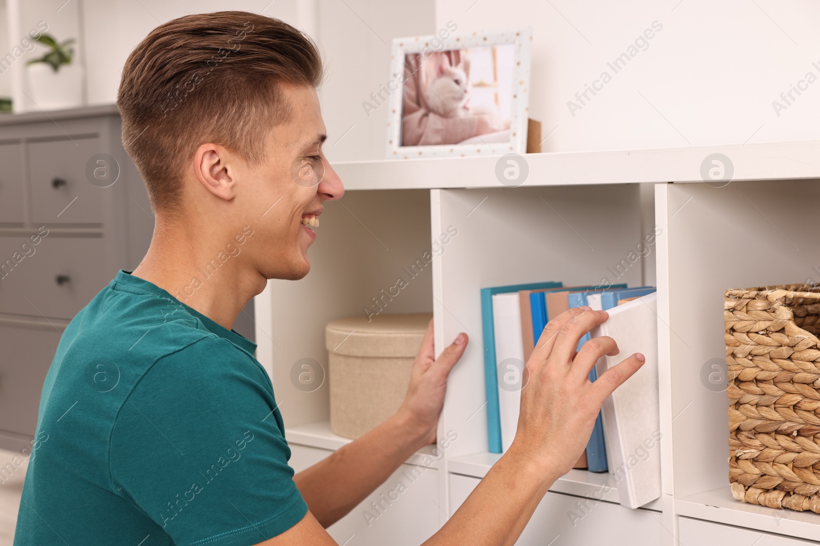 Photo of Smiling young decorator arranging books onto shelf indoors