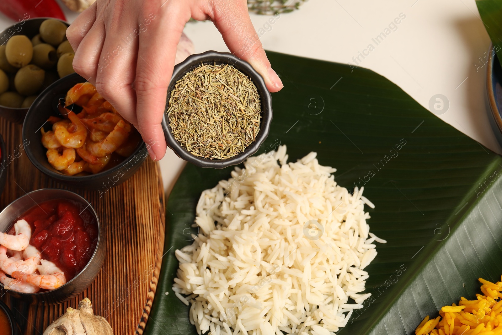 Photo of Woman holding bowl of cumin at white table with banana leaf and tasty food, closeup