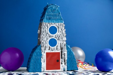 Photo of Bright pinata in shape of spaceship, balloons and confetti on table against blue background