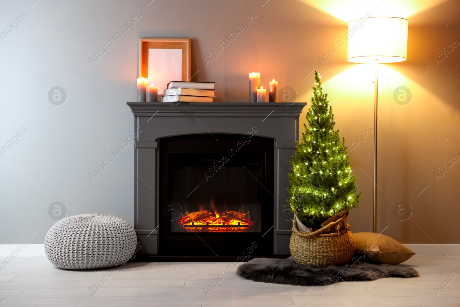 Photo of Small spruce tree in basket decorated with Christmas lights near fireplace indoors