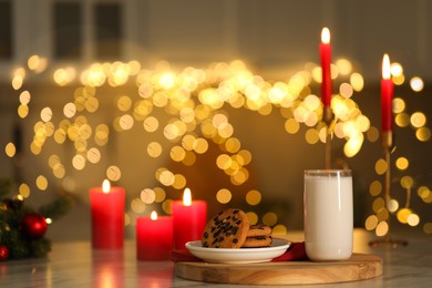 Photo of Glass of milk, tasty chocolate chip cookies and burning candles on table against blurred Christmas lights. Bokeh effect