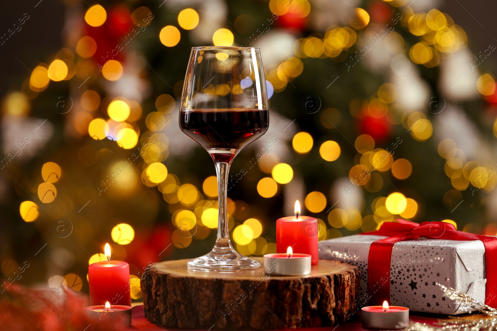 Photo of Red wine in glass, burning candles and gift box on table against blurred Christmas lights, bokeh effect