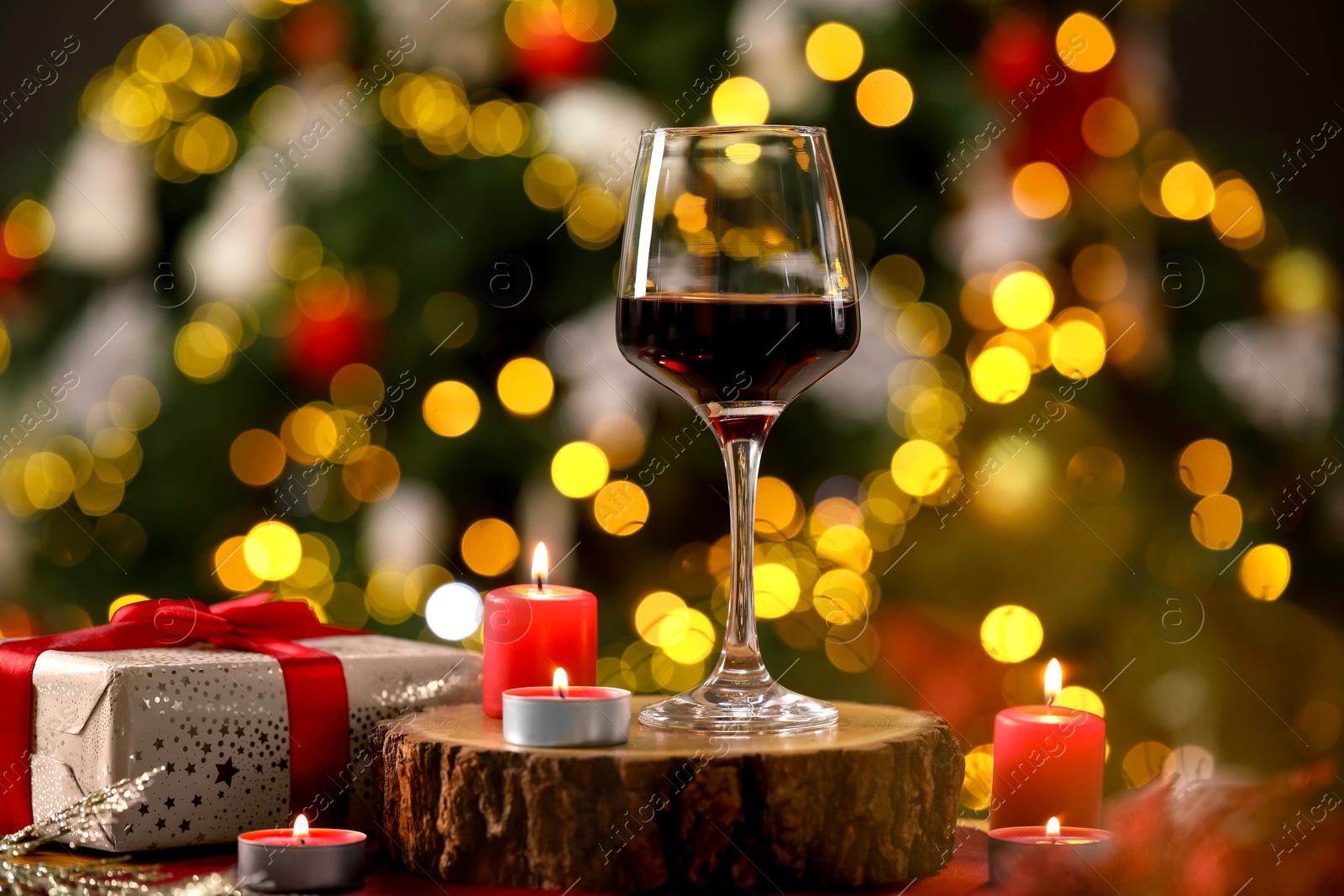 Photo of Red wine in glass, burning candles and gift box on table against blurred Christmas lights, bokeh effect