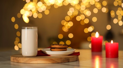 Photo of Glass of milk, tasty chocolate chip cookies and burning candles on table against blurred Christmas lights. Bokeh effect