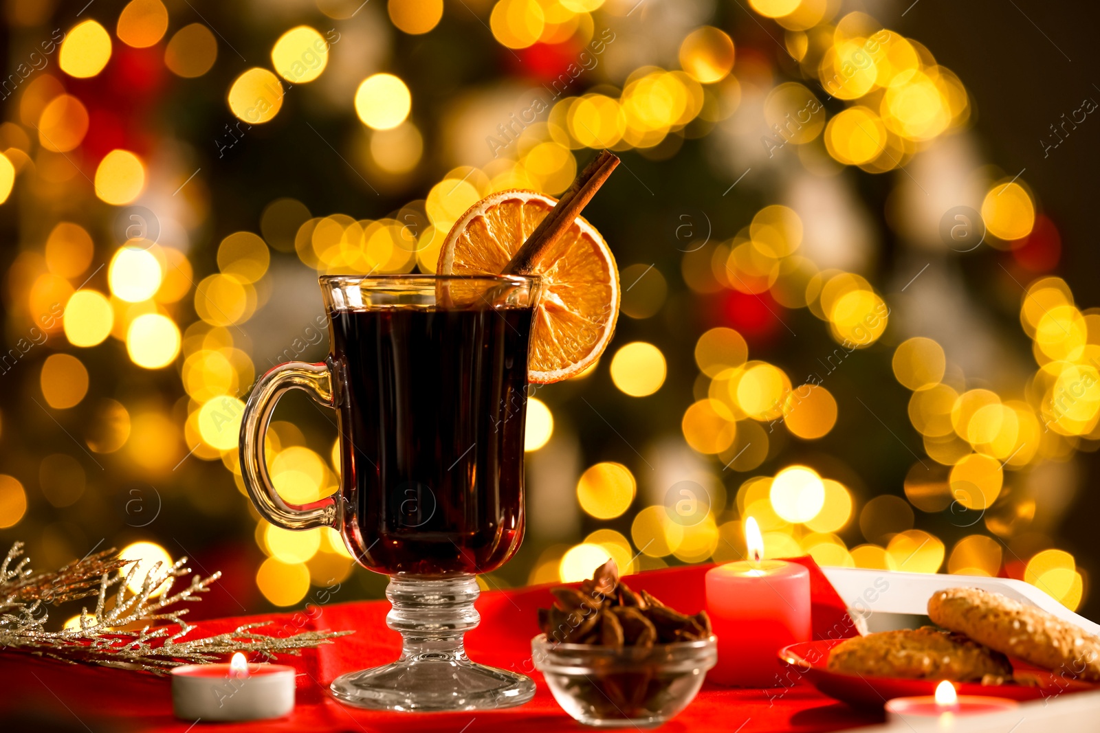 Photo of Aromatic mulled wine in glass cup, anise, cookies and burning candles on tray against blurred Christmas lights, bokeh effect