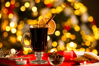 Photo of Aromatic mulled wine in glass cup, anise, cookies and burning candles on tray against blurred Christmas lights, bokeh effect