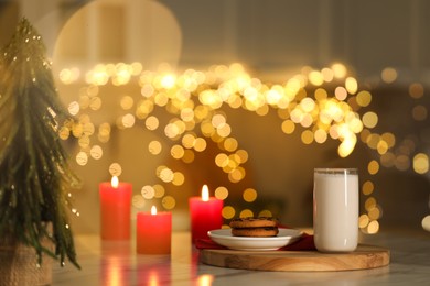 Photo of Glass of milk, tasty chocolate chip cookies and burning candles on table against blurred Christmas lights. Bokeh effect