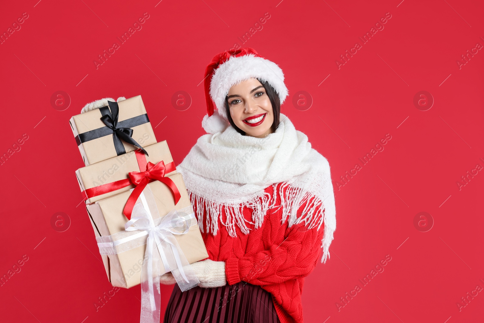 Photo of Woman in Santa hat, scarf and sweater holding Christmas gifts on red background