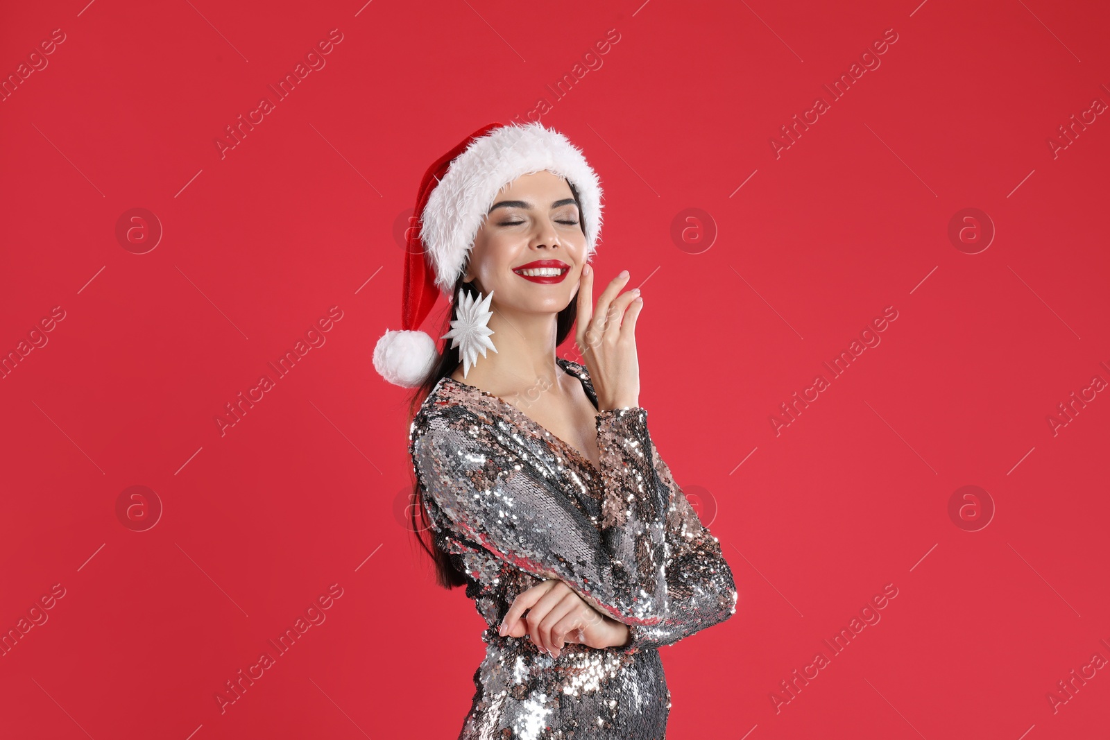 Photo of Woman in silver dress and Santa hat with snowflake earring on red background. Christmas celebration