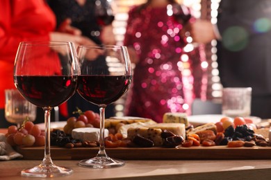 Photo of People holding glasses of wine at party, focus on wooden table with snacks and alcohol