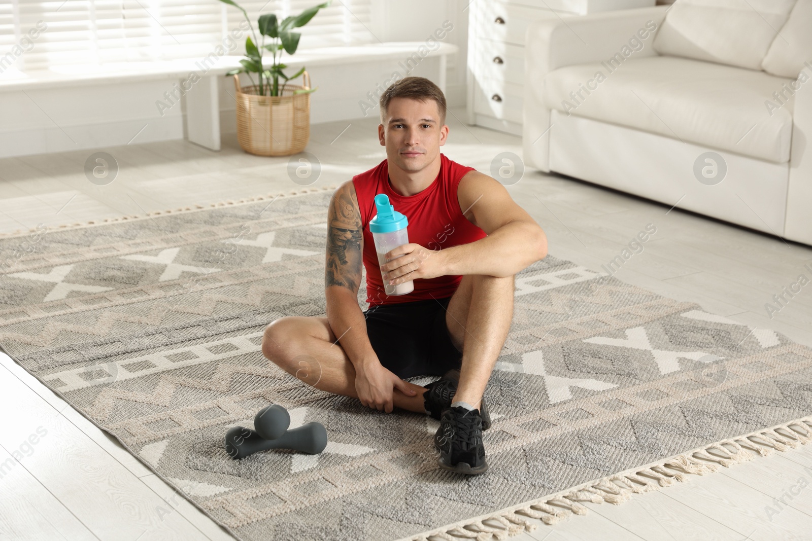 Photo of Athletic man with shaker of protein drink sitting on carpet at home