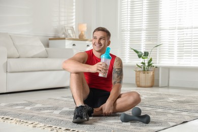 Smiling athletic man with shaker of protein drink sitting on carpet at home