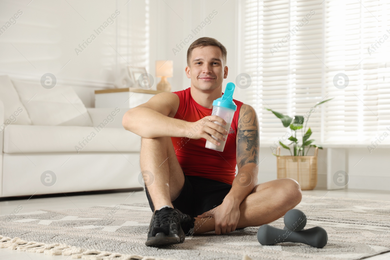 Photo of Athletic man with shaker of protein drink sitting on carpet at home