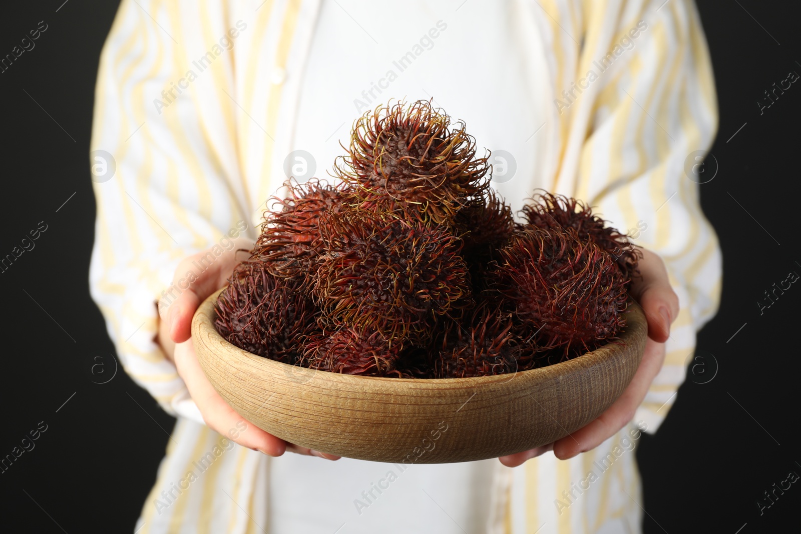 Photo of Woman holding fresh rambutans in bowl on black background, closeup