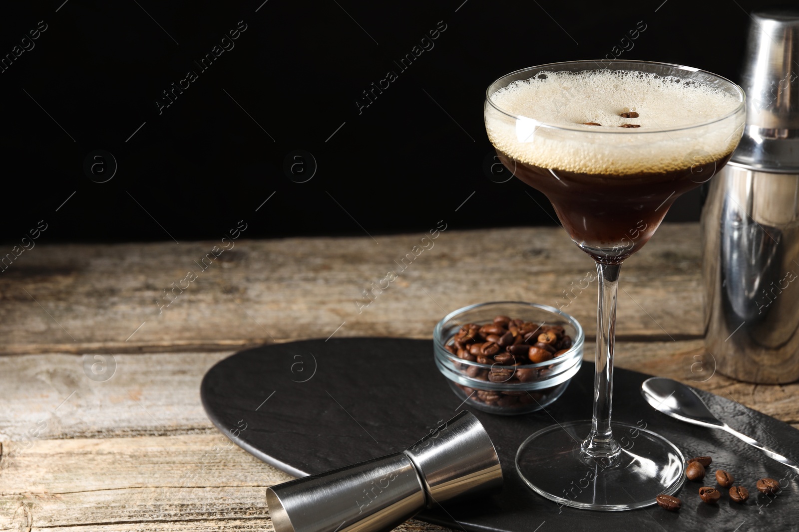 Photo of Glass of delicious espresso martini, jigger and coffee beans on wooden table against black background, closeup. Space for text