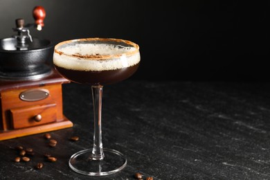 Photo of Glass of delicious espresso martini, manual grinder and coffee beans on dark textured table, closeup. Space for text