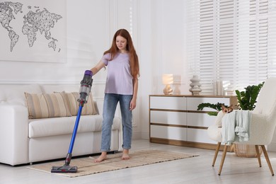 Photo of Teenage girl cleaning rug with cordless vacuum cleaner and her cute Chihuahua dog sitting in armchair at home