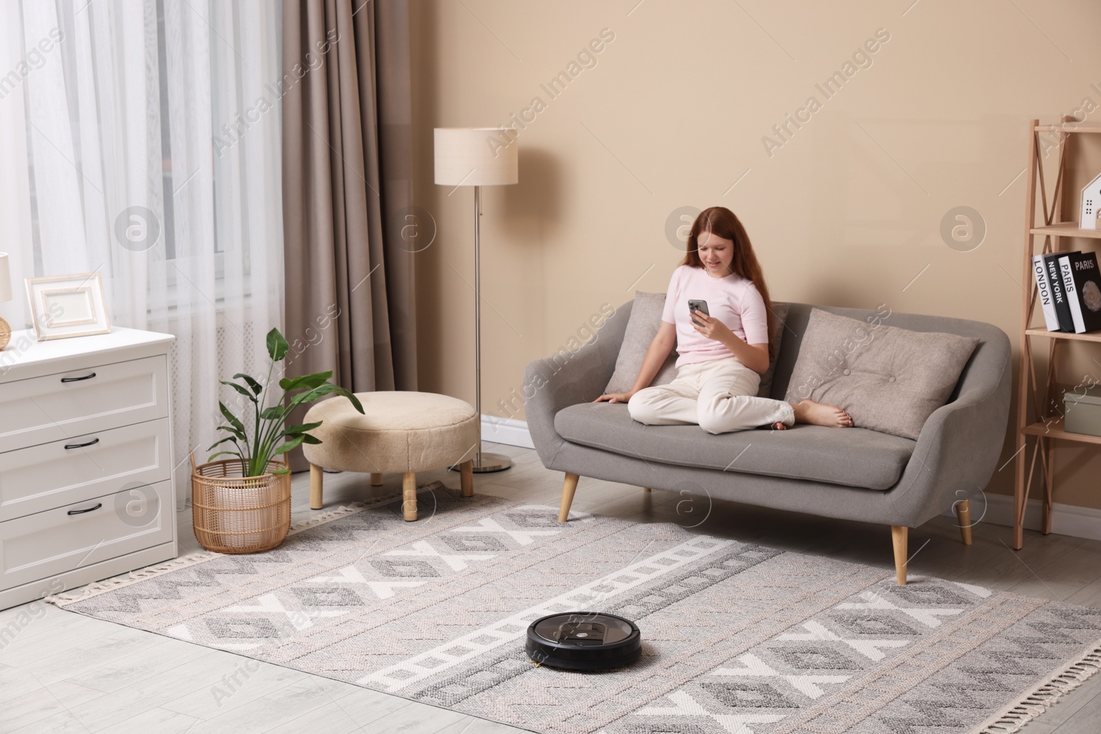 Photo of Teenage girl with smartphone while robotic vacuum cleaner vacuuming rug at home