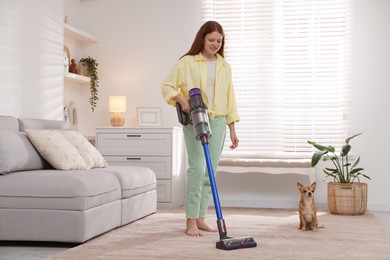 Photo of Teenage girl cleaning rug with cordless vacuum cleaner and her cute Chihuahua dog sitting on floor at home