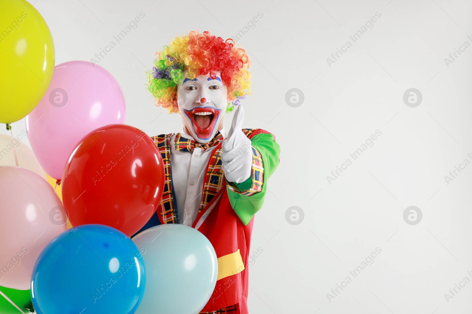 Photo of Happy clown with colorful balloons showing thumbs up on light background, space for text