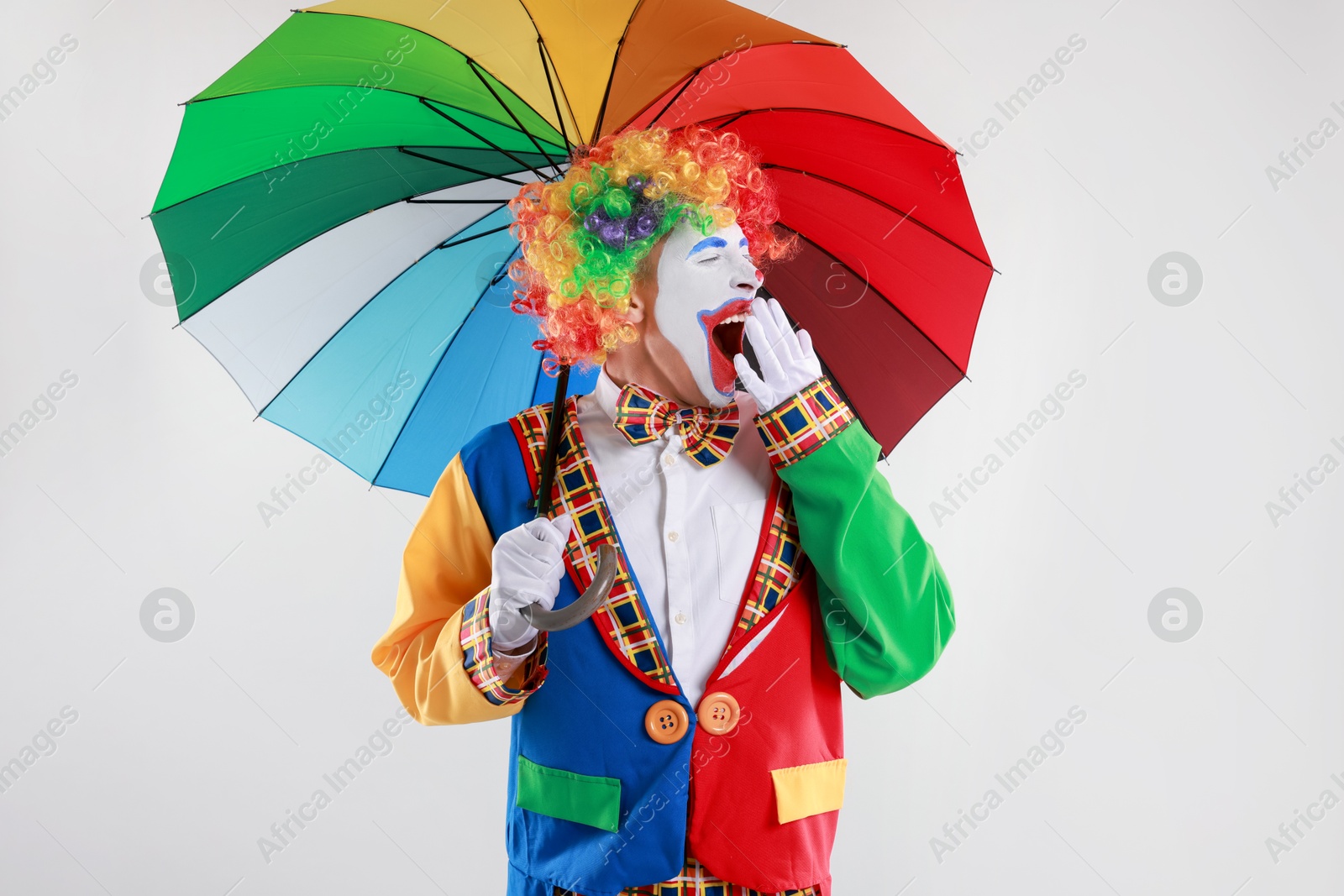 Photo of Clown with colorful umbrella yawning on light background