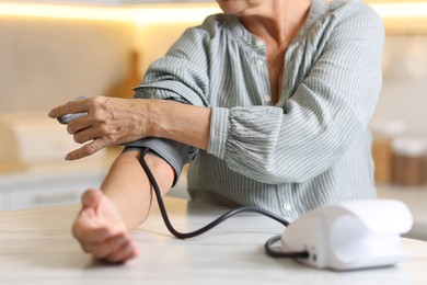 Photo of Senior woman measuring blood pressure at table in kitchen, closeup