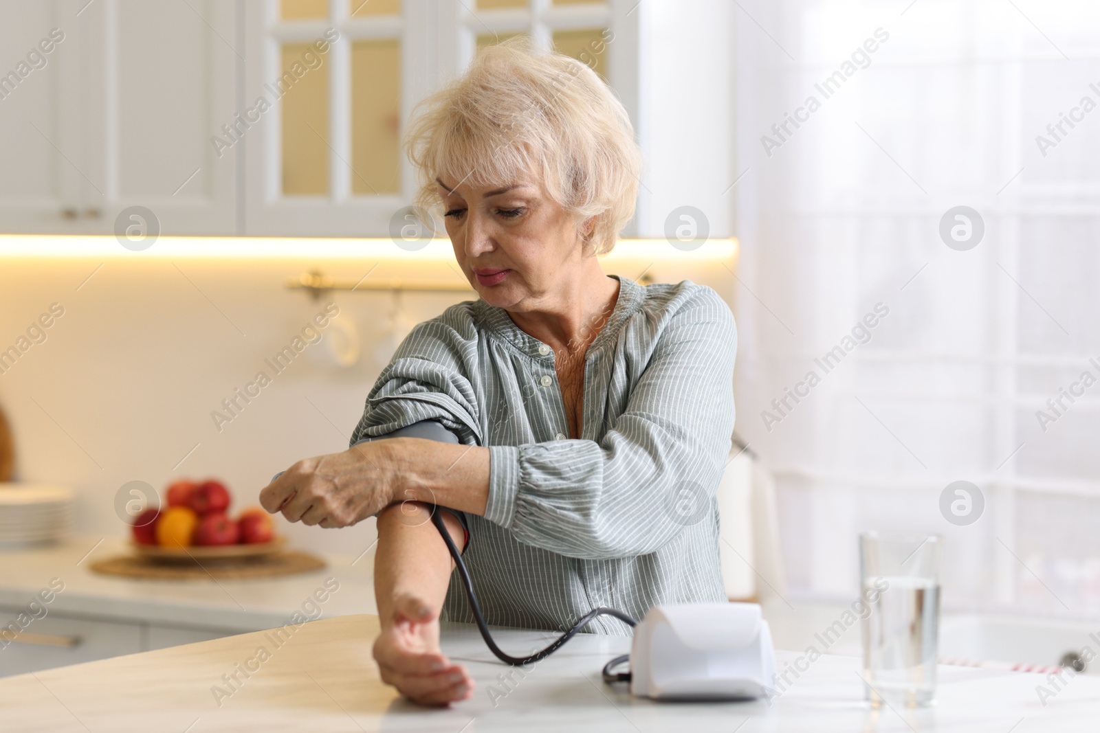 Photo of Senior woman measuring blood pressure at table in kitchen